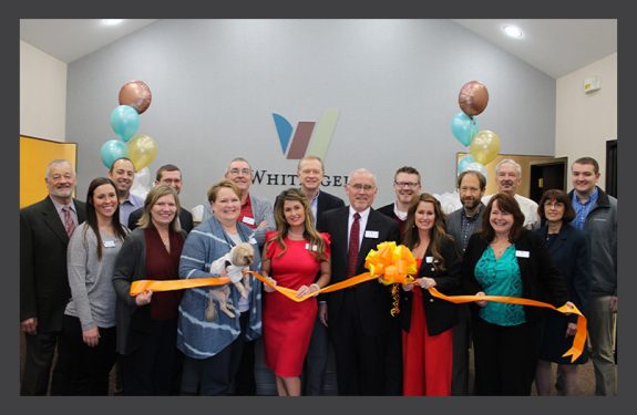 Various Whitinger & Company employees and member-owners, smiling and holding a large yellow ribbon tied in a bow.